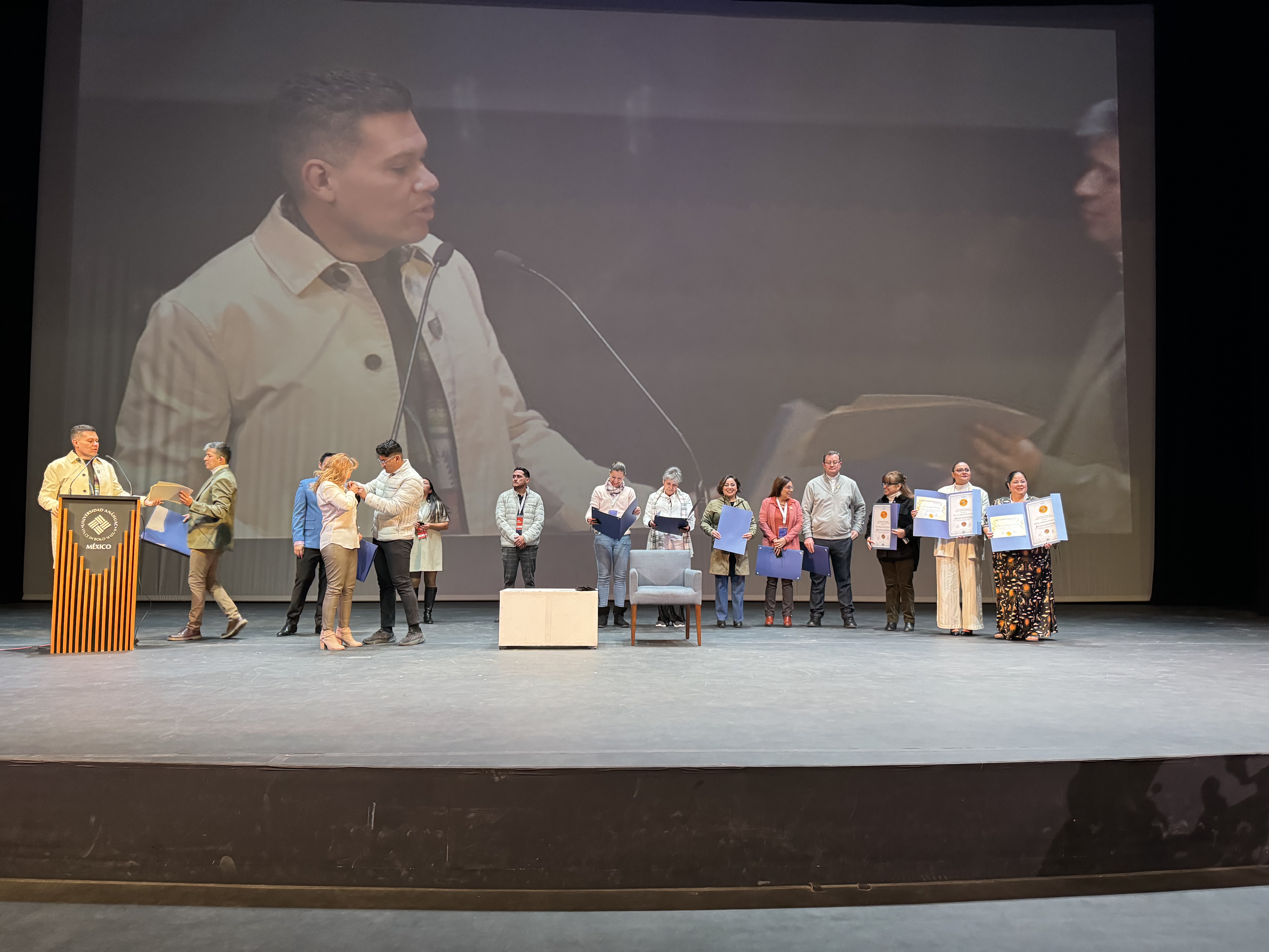 Oscar Rivas en el podio de la Universidad Anáhuac entregando reconocimientos, con su imagen proyectada en pantalla